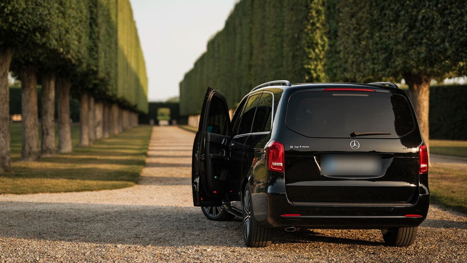 A black Mercedes V-Class with its side door open on the gravel drive of a Loire Valley estate, between clipped hedge avenues