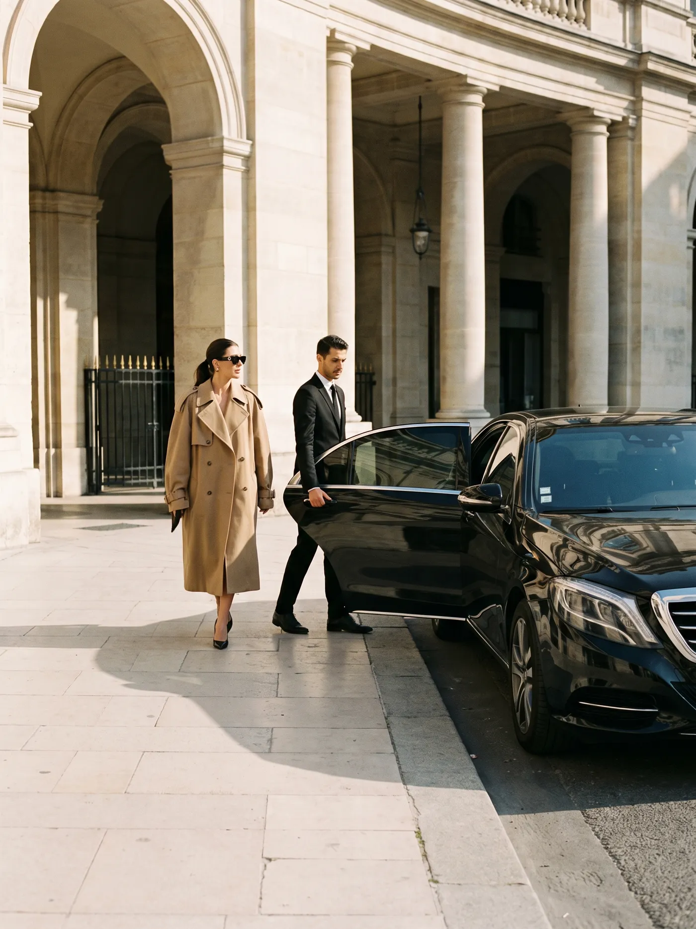 A woman in a camel coat and sunglasses stepping towards a black Mercedes S-Class under a Paris stone colonnade, the chauffeur holding the door