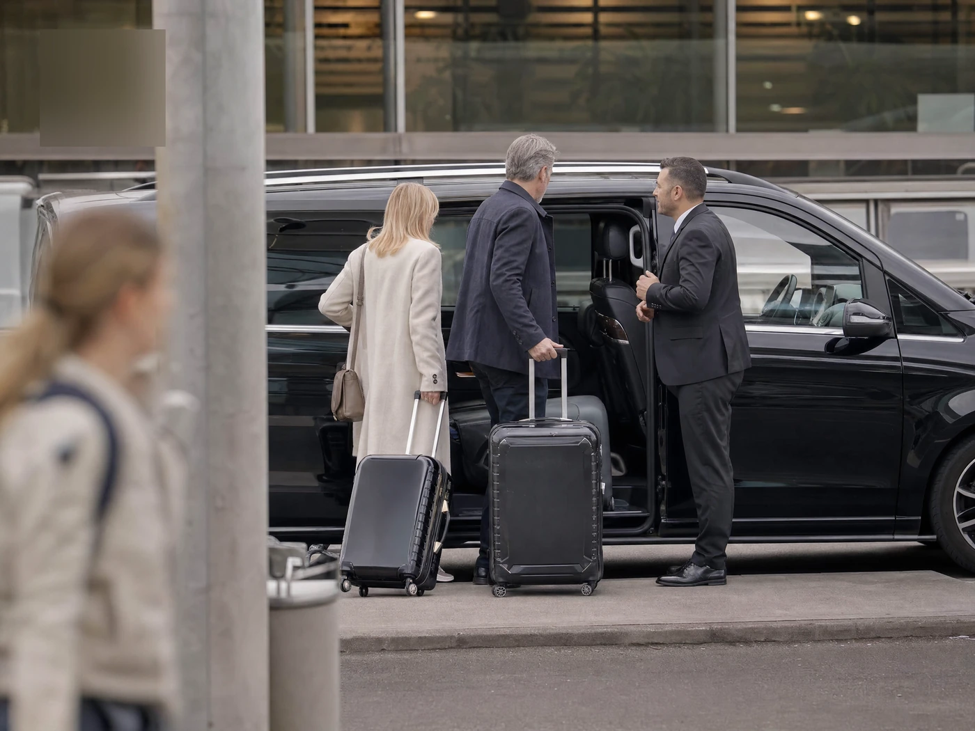 Chauffeur greeting a couple with their suitcases beside a Mercedes V-Class outside an airport terminal