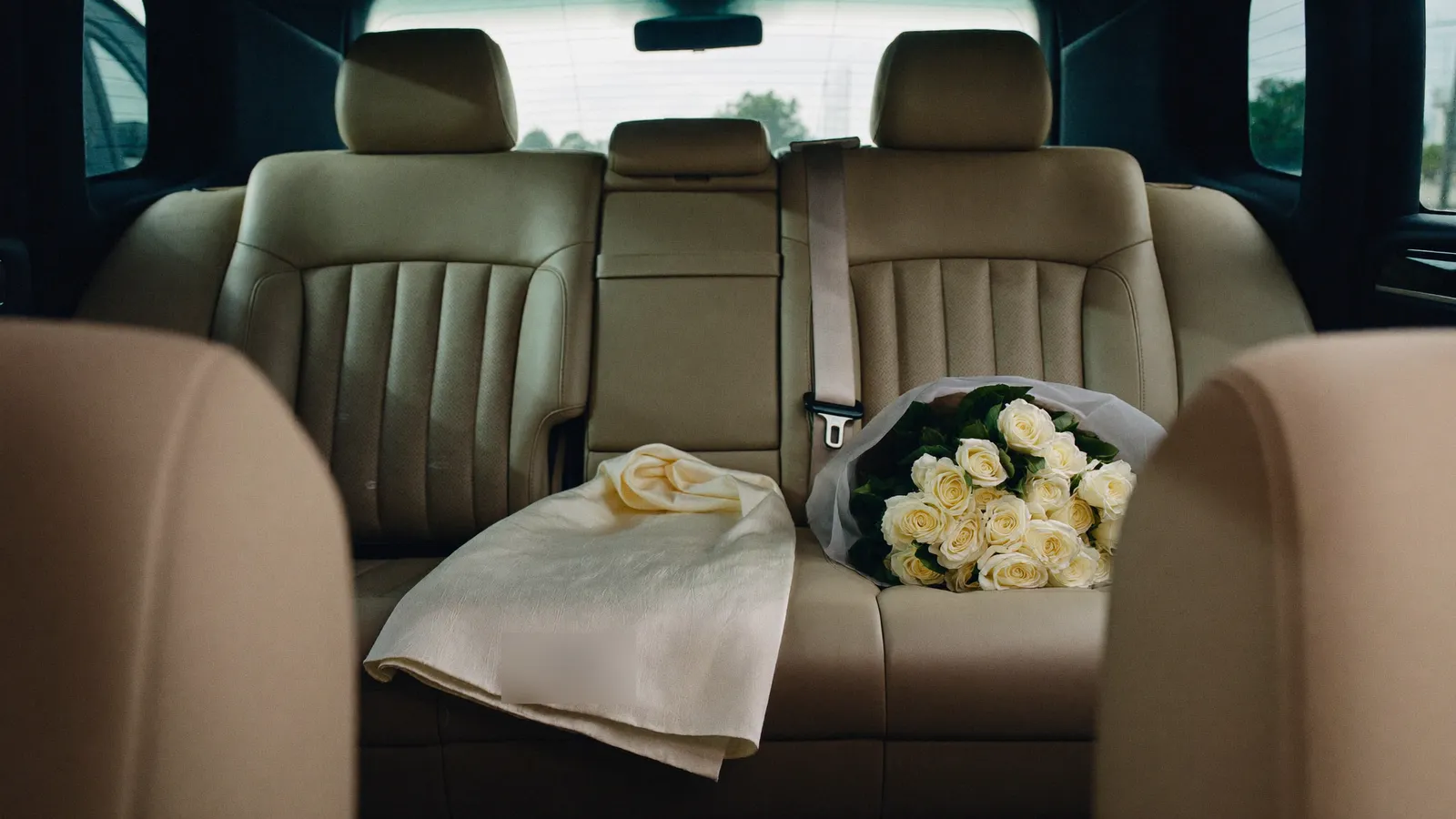 Cream leather rear bench of a chauffeured Mercedes, with a bouquet of white roses and a folded garment laid on the seat